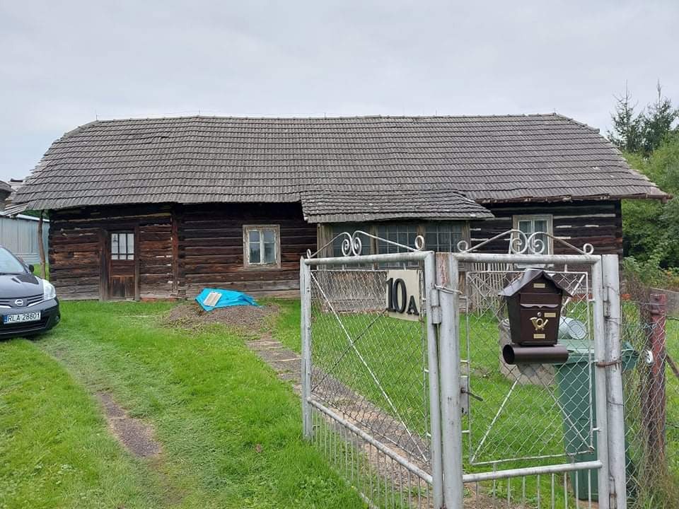 Stary, drewniany dom z ciemną, spadzistą dachówką, widoczny zza metalowej furtki z numerem 10A, obok skrzynka pocztowa i zaparkowany samochód.