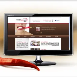Monitor wyświetla stronę internetową dentysty z logo i zdjęciem kobiety trzymającej papryczkę chili w ustach, obok monitora leży prawdziwa czerwona papryka chili.