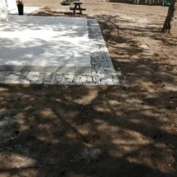 Wykończony taras z jasnoszarej płyty obrzeżony kostką granitową, z widocznym domkiem dla dzieci w tle.