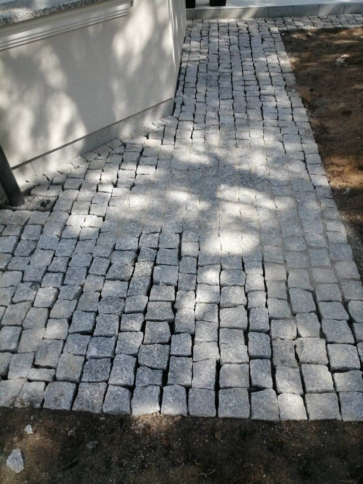 Ścieżka z szarej kostki granitowej ułożona przy ścianie budynku, z widocznymi cieniami drzew.