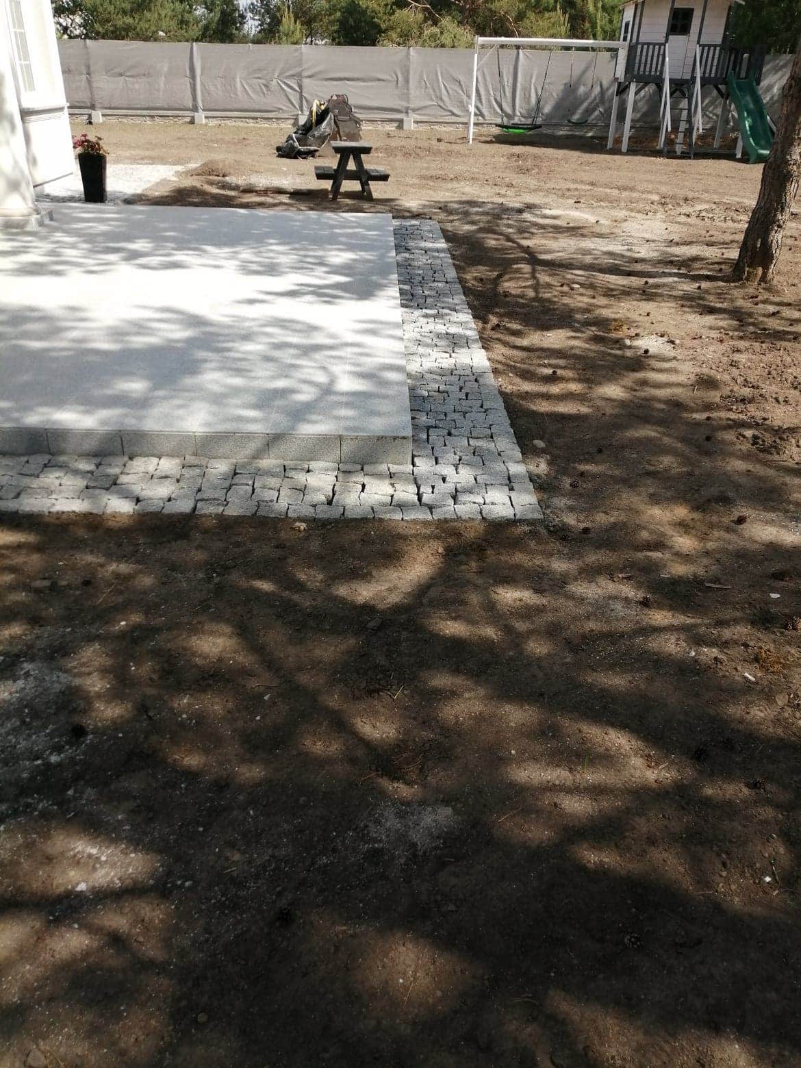 Wykończony taras z jasnoszarej płyty obrzeżony kostką granitową, z widocznym domkiem dla dzieci w tle.