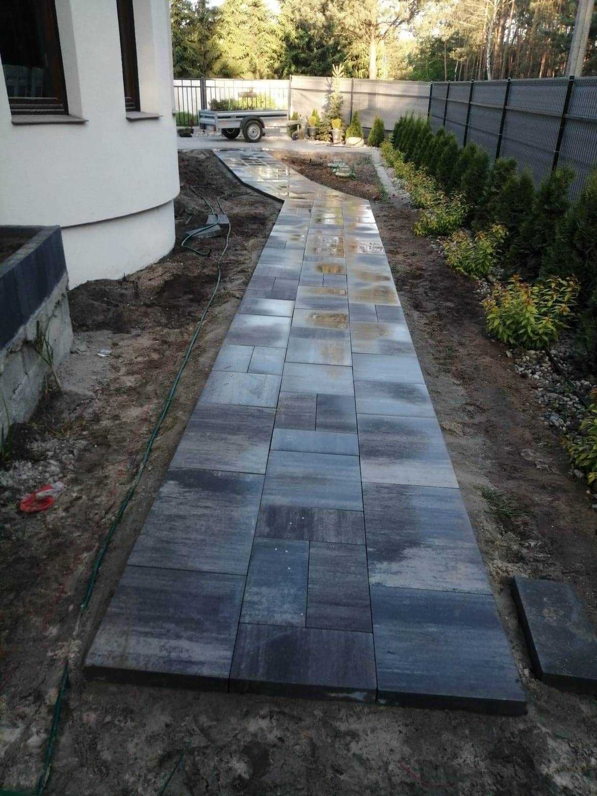 Świeżo ułożona, długa ścieżka z szarych, prostokątnych kostek brukowych prowadząca wzdłuż białego budynku i ogrodzenia, widoczne ślady wilgoci na powierzchni kostki, trawa i krzewy po prawej stronie.