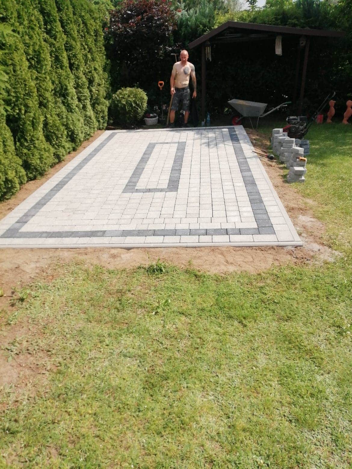 Ułożona kostka brukowa w ogrodzie z dekoracyjnym wzorem obramowania i częściowo ułożonymi kostkami obok, mężczyzna z łopatą w tle.