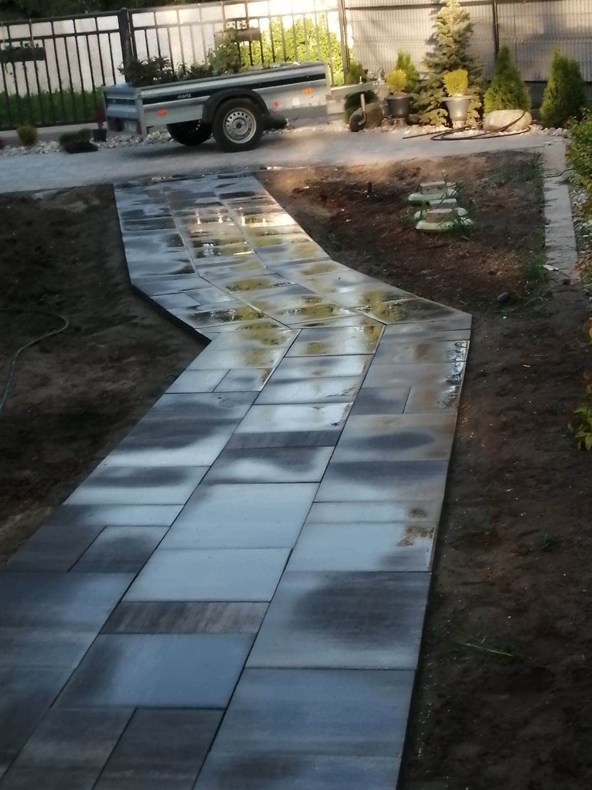 Świeżo ułożony, wilgotny chodnik z prostokątnej kostki brukowej w odcieniach szarości, wijący się wzdłuż rabaty z zielenią, z przyczepką samochodową w tle.