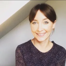 Uśmiechnięta kobieta z brązowymi włosami i niebieskimi oczami, ubrana w bluzkę w drobny wzór, pozuje na tle jasnej ściany i fragmentu skośnego sufitu.