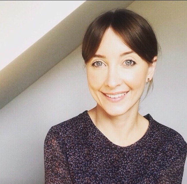 Uśmiechnięta kobieta z brązowymi włosami i niebieskimi oczami, ubrana w bluzkę w drobny wzór, pozuje na tle jasnej ściany i fragmentu skośnego sufitu.