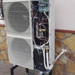 Otwarta obudowa zewnętrznej jednostki pompy ciepła na czarnym metalowym stelażu, widoczne wewnętrzne komponenty, rury miedziane i kable, na tle ściany budynku wykończonej kamieniem.