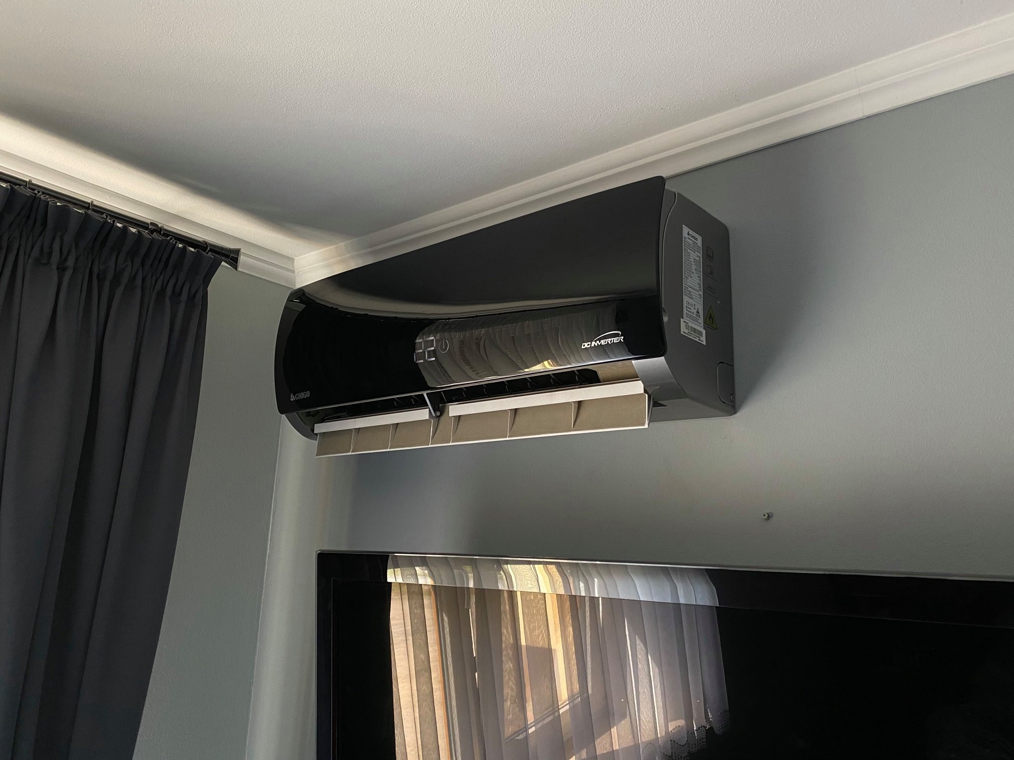Czarny, błyszczący klimatyzator ścienny marki Chigo z wyświetlaczem temperatury 22 stopnie Celsjusza, zamontowany nad telewizorem w szarym pokoju z zasłonami.