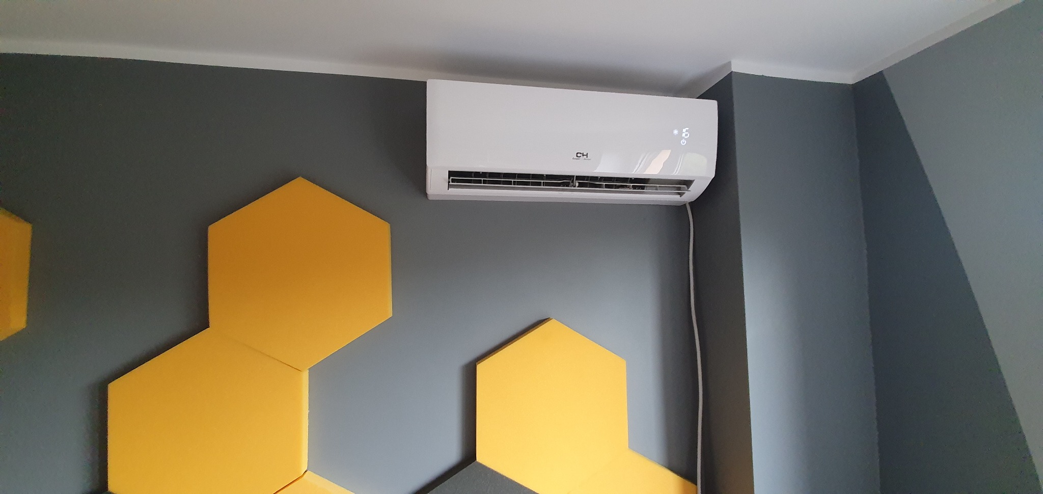 Biały klimatyzator na szarej ścianie z dekoracyjnymi, żółtymi panelami w kształcie plastra miodu; widoczny kabel zasilający.