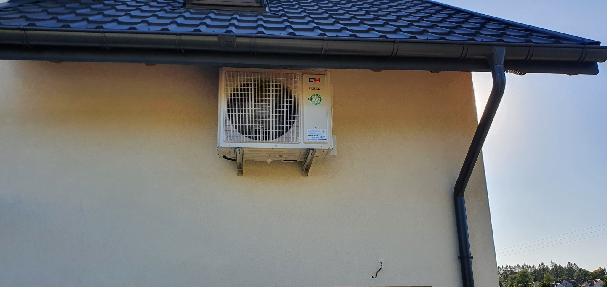 Zewnętrzna jednostka klimatyzacji marki CH zamontowana na elewacji domu, widoczne metalowe wsporniki i fragment dachu z orynnowaniem.