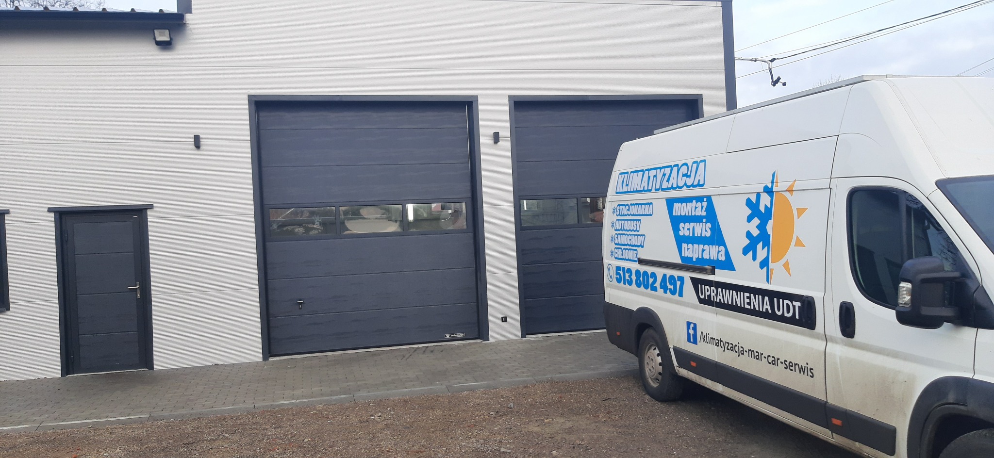 Biała furgonetka z niebiesko-żółtym logo firmy zajmującej się klimatyzacją, zaparkowana przed budynkiem z dwiema bramami garażowymi i drzwiami wejściowymi.