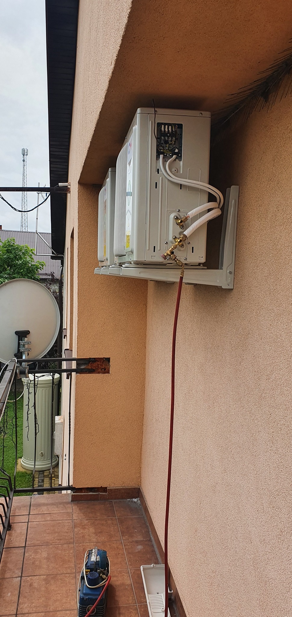 Instalacja zewnętrznych jednostek klimatyzacji na balkonie budynku o beżowej elewacji, widoczne białe rury i przewody, na podłodze balkonu narzędzia i pojemnik.