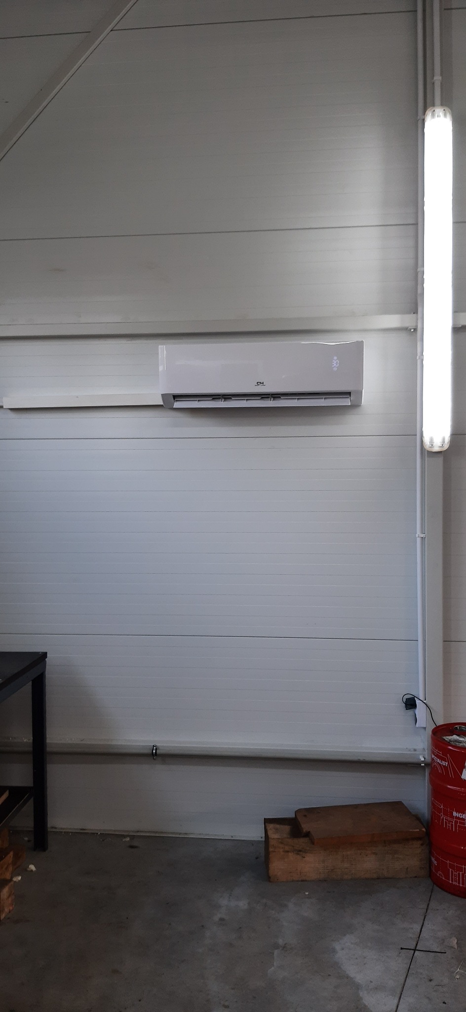 Biały klimatyzator zamontowany na ścianie z panelami, widoczny element instalacji elektrycznej oraz metalowa beczka i drewniana skrzynka na podłodze.