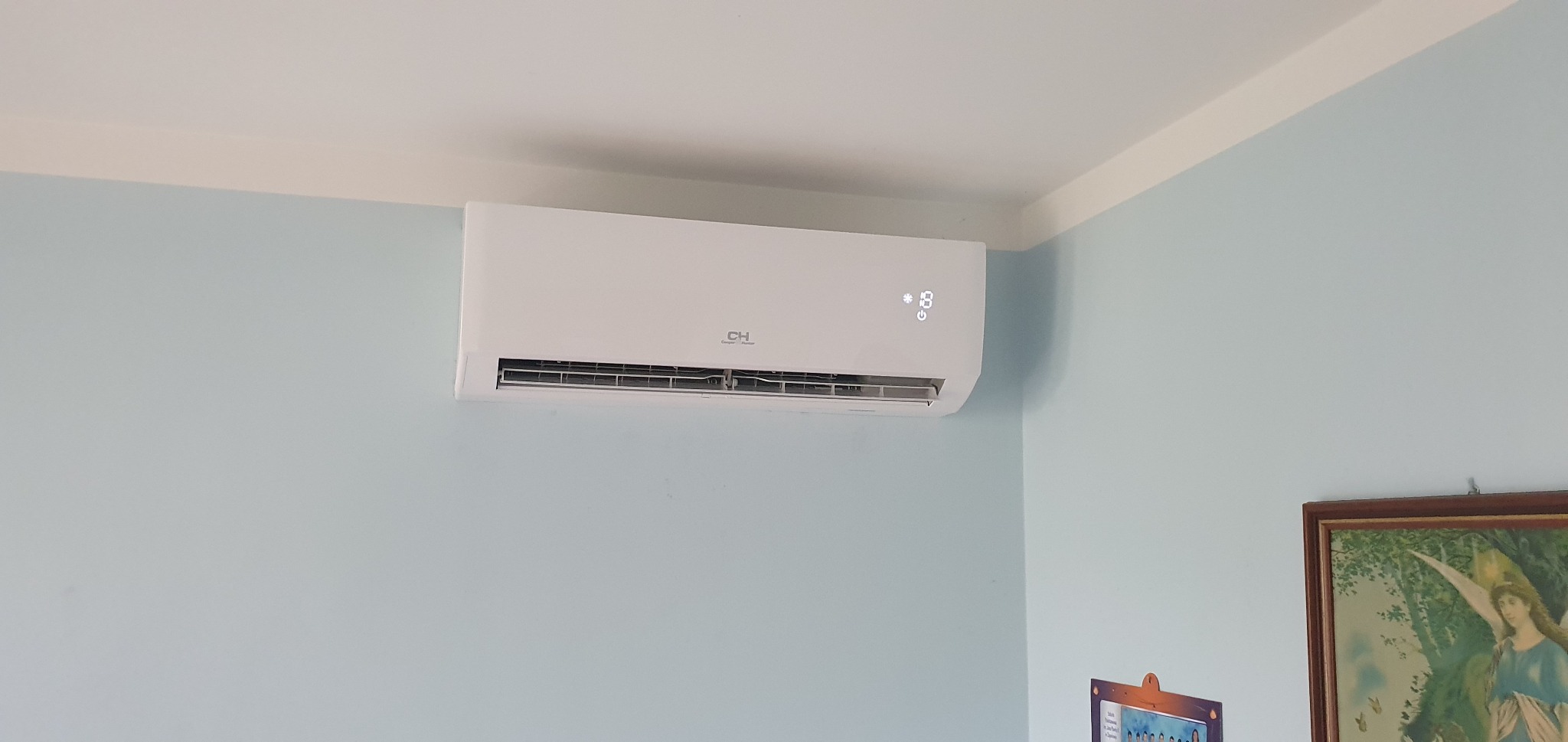 Biała jednostka klimatyzacyjna marki CH zamontowana na jasnoniebieskiej ścianie w rogu pokoju, z widocznym wyświetlaczem temperatury i obrazem w ramce w tle.