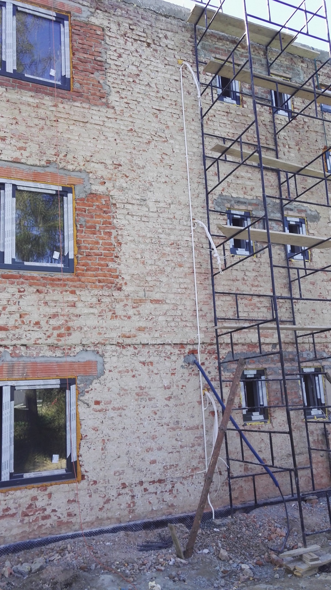 Okablowanie poprowadzone po elewacji budynku z cegły, obok rusztowanie, przygotowane do montażu urządzeń klimatyzacyjnych po remoncie bez kucia w ścianach.