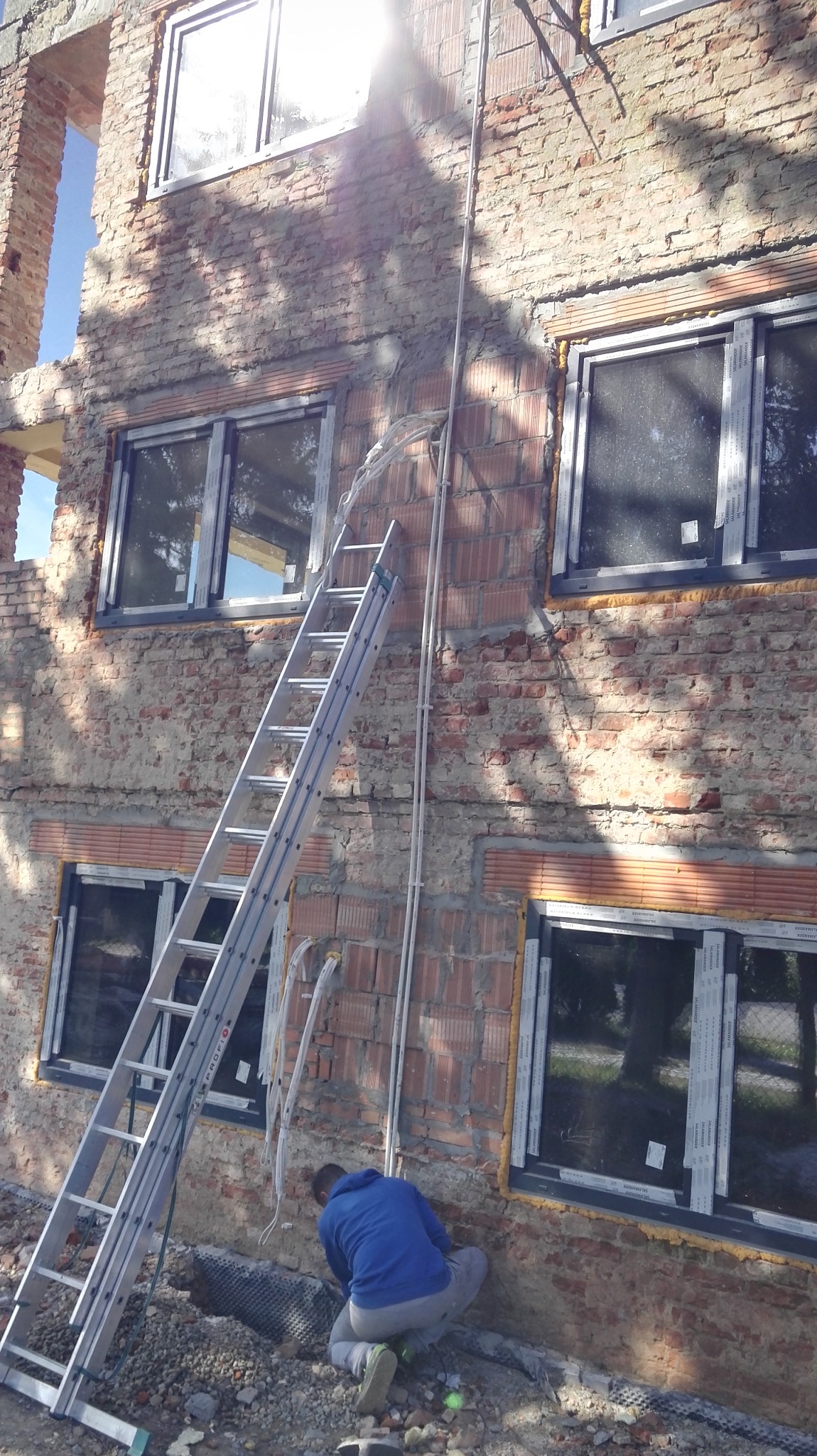 Montaż instalacji przewodów na zewnętrznej ścianie budynku z cegły, widoczna drabina oparta o ścianę, pracownik w niebieskiej bluzie w trakcie pracy przy instalacji.