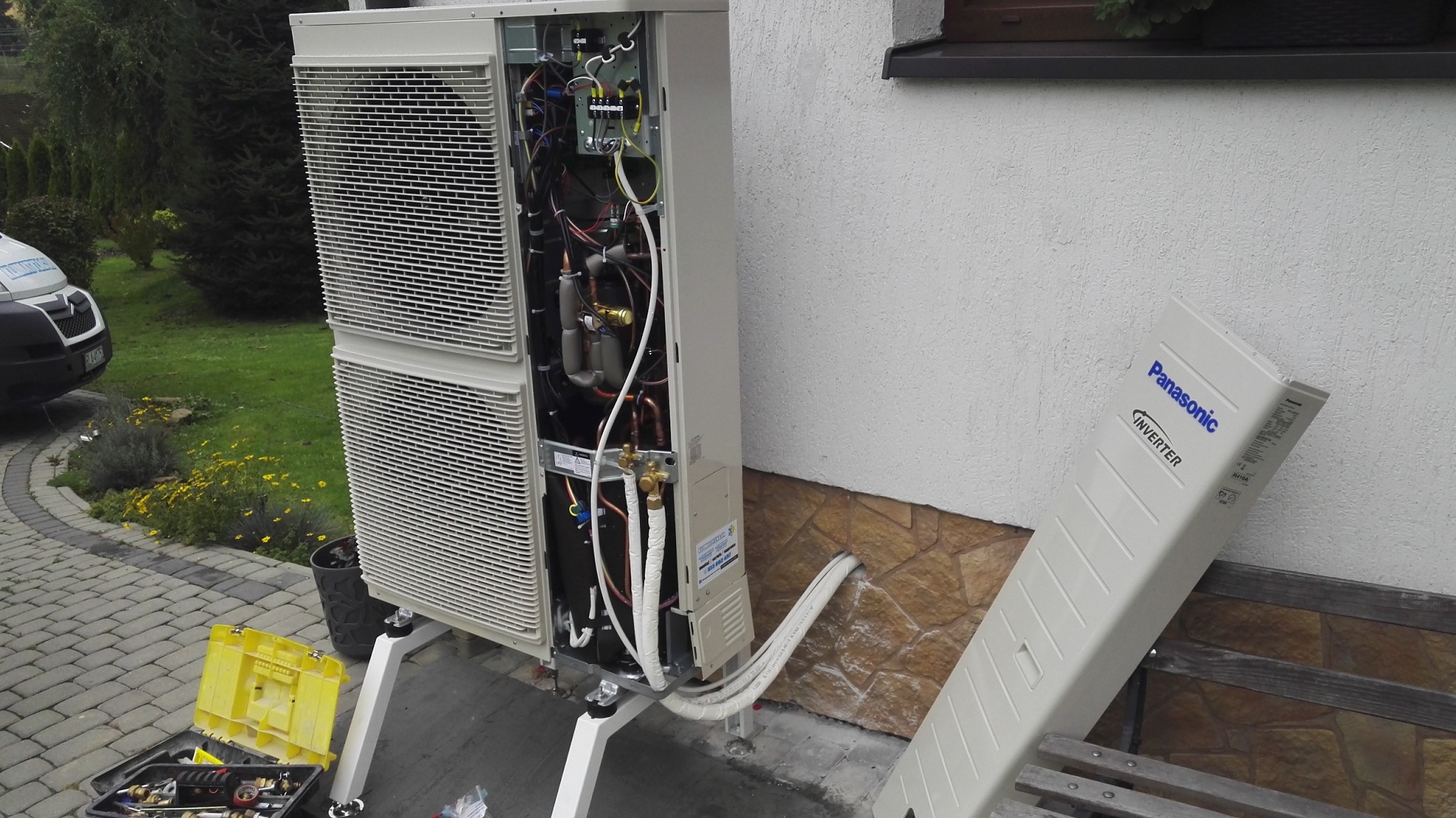 Zewnętrzna jednostka pompy ciepła Panasonic Inverter w trakcie montażu, widoczne podłączenia przewodów, narzędzia w żółtej skrzynce obok, częściowo zdjęta obudowa, tło stanowi budynek i zieleń.
