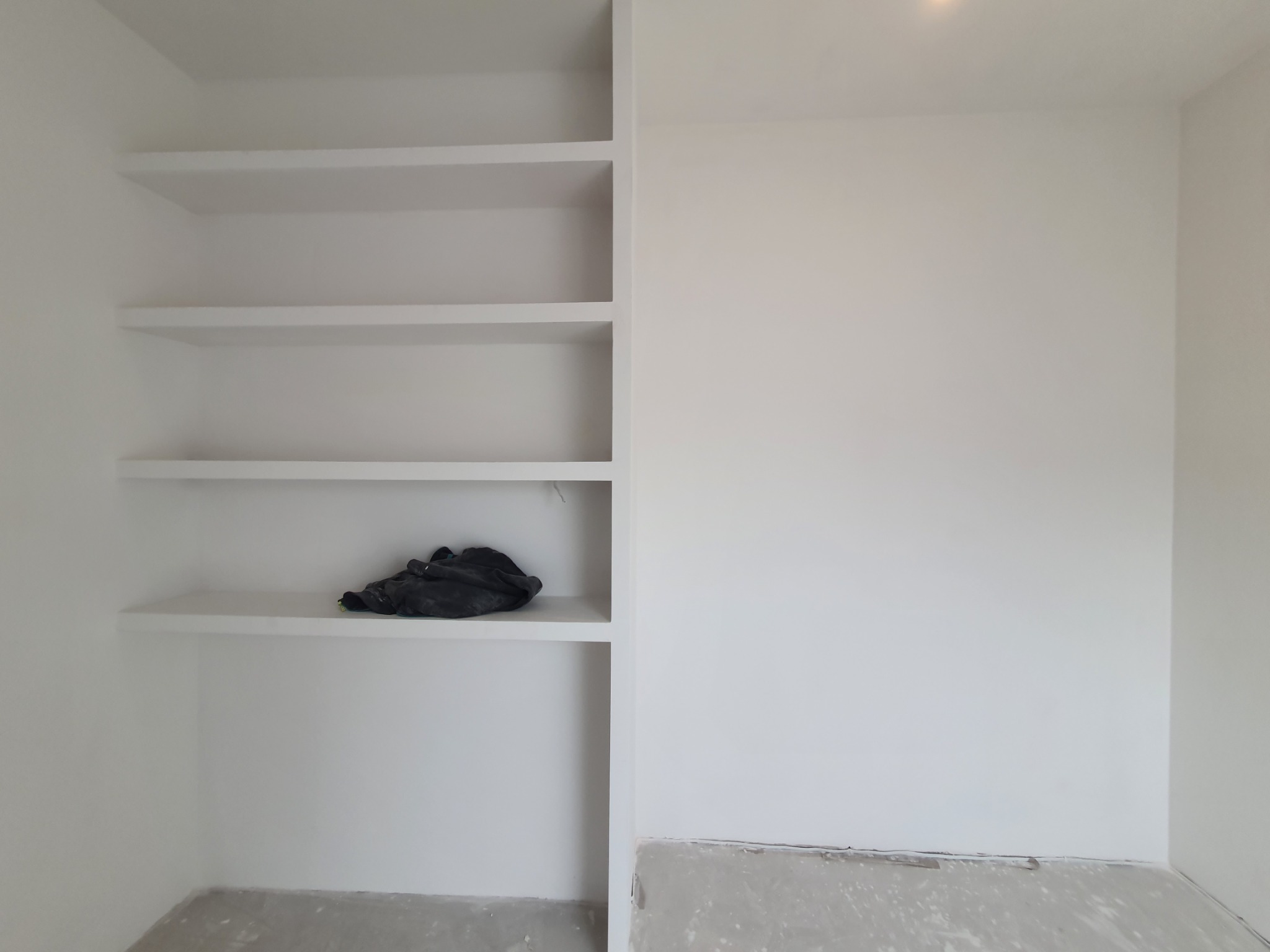 Białe, wbudowane półki w ścianie po remoncie w Warszawie, z czarnym workiem na jednej z półek i widocznymi śladami farby na podłodze.