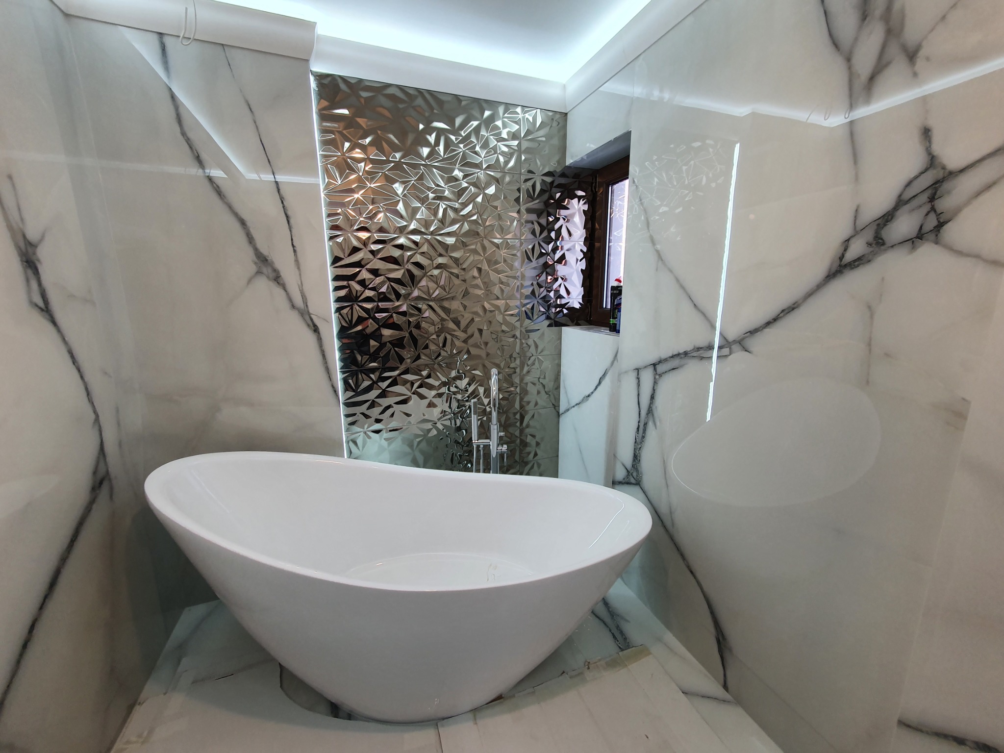 Biała, wolnostojąca wanna w łazience wykończonej marmurowymi płytami i dekoracyjnymi, srebrnymi panelami 3D na ścianie za wanną, z oknem w drewnianej ramie.