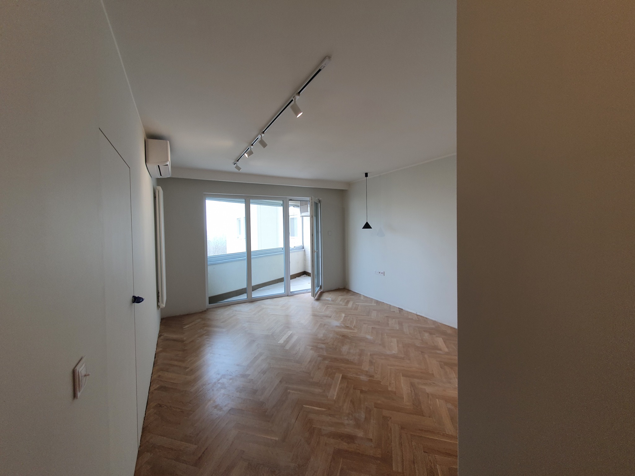 Pusty pokój po odświeżeniu z parkietem ułożonym w jodełkę, białymi ścianami, przesuwnymi drzwiami balkonowymi i nowoczesnym oświetleniem szynowym.