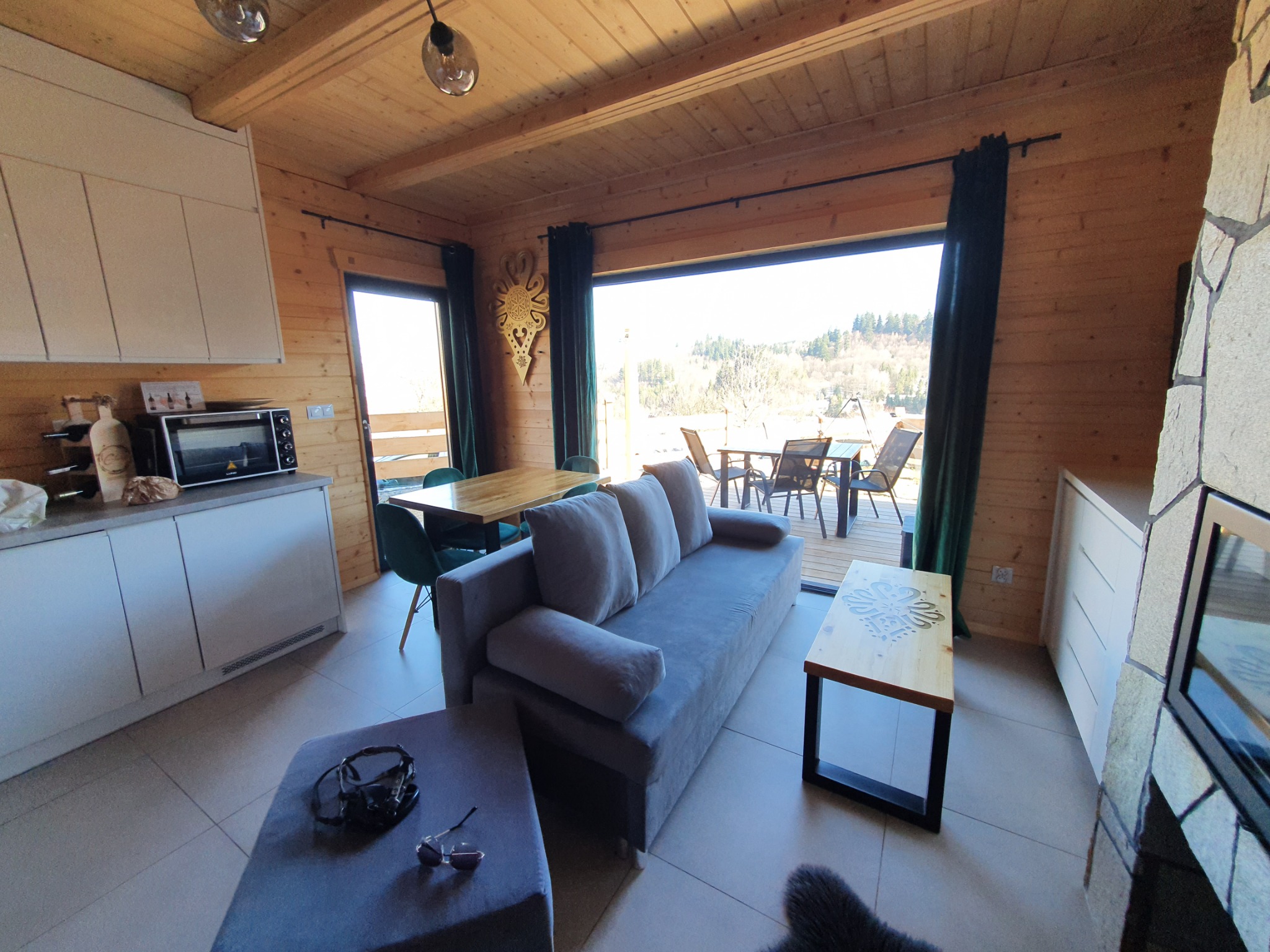 Jasne wnętrze drewnianego domu z widokiem na zewnątrz, szary narożnik, drewniane meble, dekoracyjny zegar na ścianie i kamienny kominek.