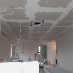Wnętrze w trakcie prac wykończeniowych: sufit z płyt gipsowo-kartonowych, częściowo zaszpachlowany, widoczne kable, drabiny i narzędzia na podłodze.