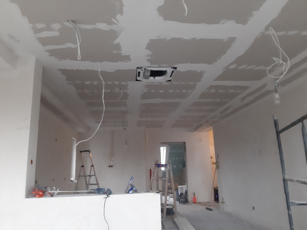 Wnętrze w trakcie prac wykończeniowych: sufit z płyt gipsowo-kartonowych, częściowo zaszpachlowany, widoczne kable, drabiny i narzędzia na podłodze.