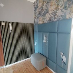 AMB Arkadiusz Kurowski - Nowoczesne wykończenie wnętrza z drewnianą podłogą, ścianą z pionowych lameli, wzorzystą tapetą i panelem tapicerowanym w kolorze błękitnym z motywem żurawi, uzupełnione szarą pufą.