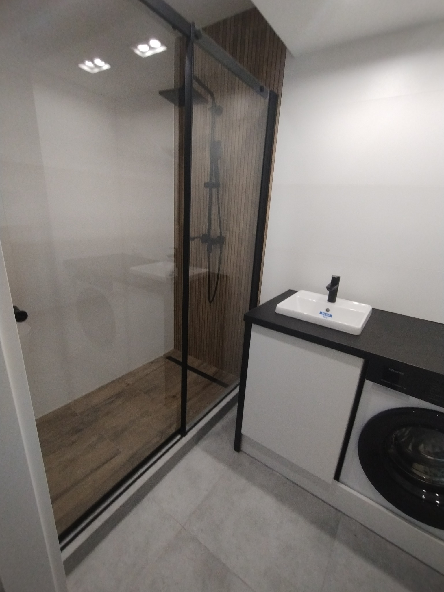 Nowoczesna łazienka z kabiną prysznicową typu walk-in z czarnymi profilami, drewnopodobnymi płytkami na ścianie i podłodze w kabinie, umywalką nablatową z czarną armaturą oraz pralką pod blatem.