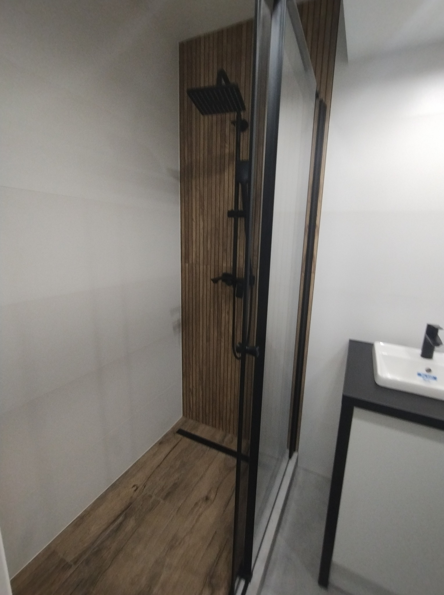 Nowoczesna łazienka z kabiną prysznicową typu walk-in z czarną armaturą, drewnopodobną okładziną ściany oraz umywalką nablatową na czarnym blacie.