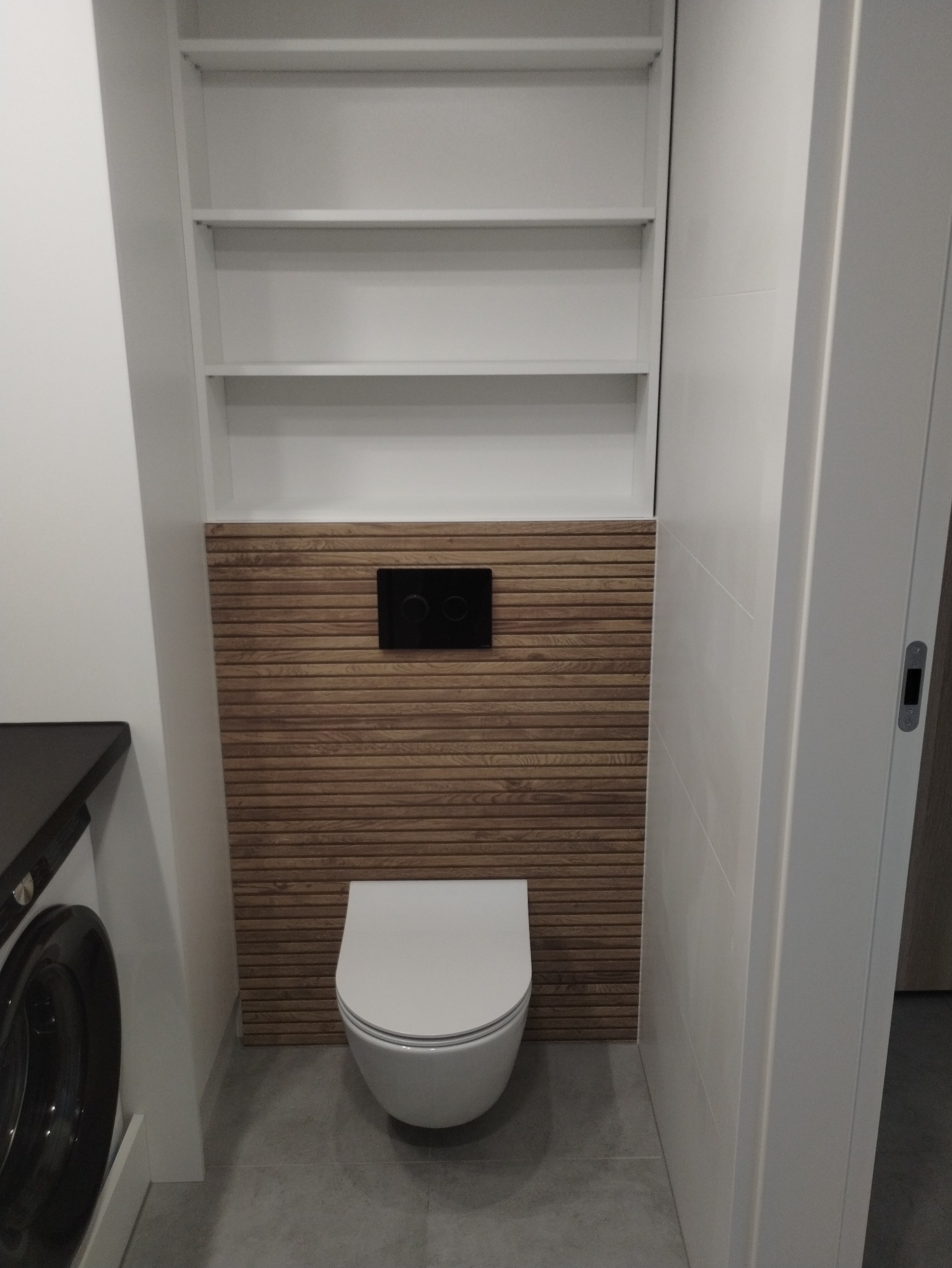 Wisząca toaleta z czarnym panelem sterującym na ścianie z drewnopodobnych paneli, nad toaletą białe półki, obok pralka.