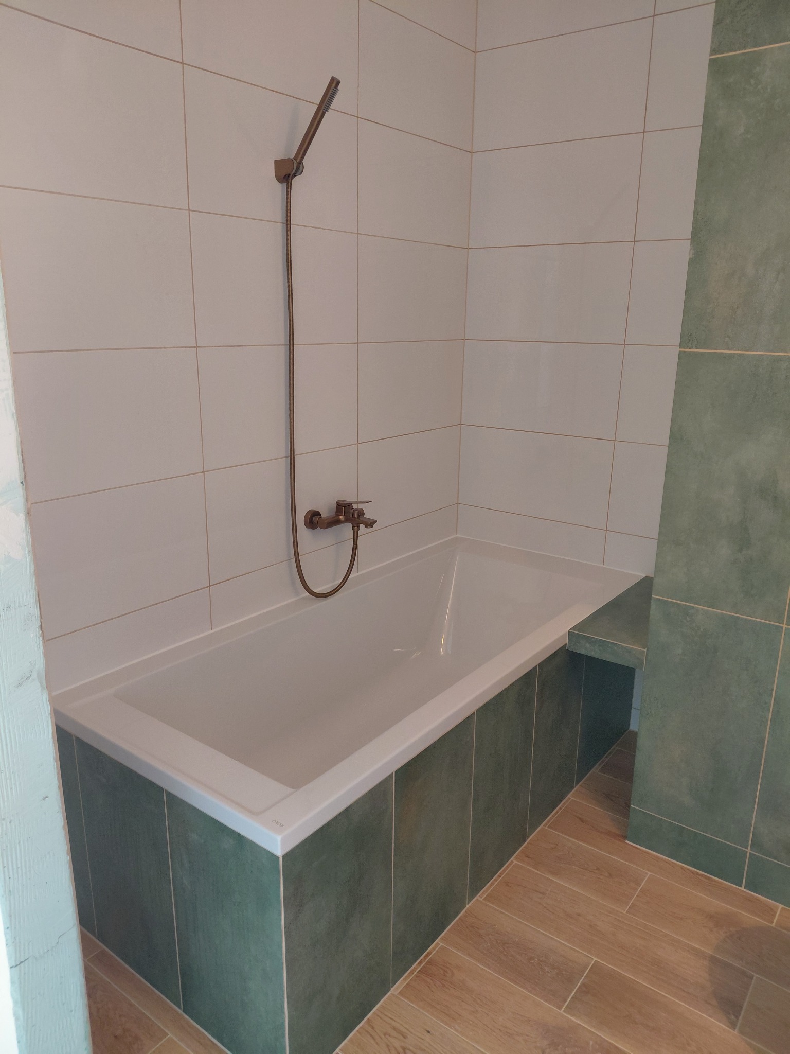 Wykończona łazienka: biała wanna obudowana zielonymi płytkami, bateria w stylu retro, jasne płytki na ścianach, podłoga imitująca drewno. Ukończona realizacja.