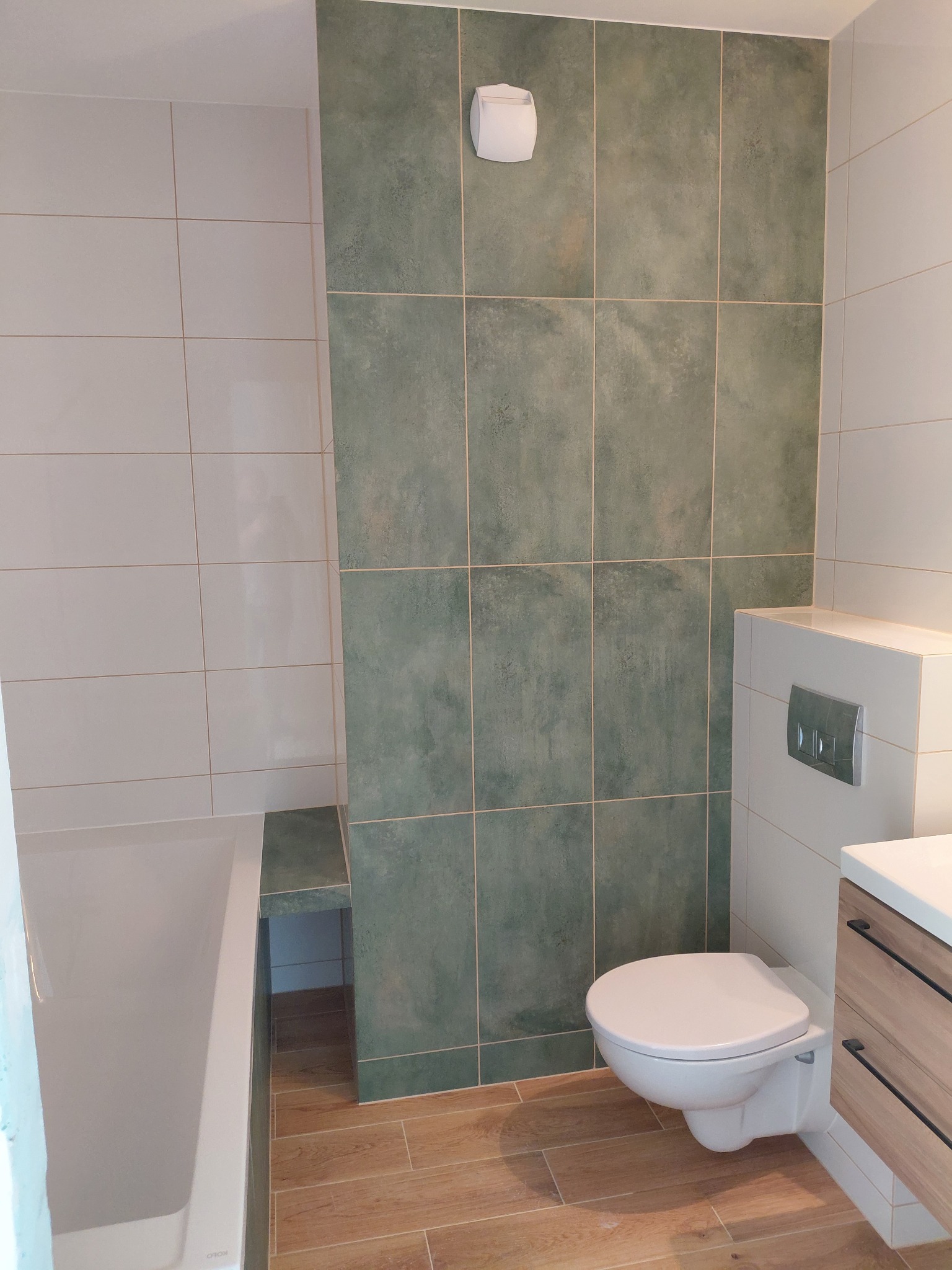 Wykończona łazienka z wanną, toaletą podwieszaną i umywalką z szafką. Ściany wyłożone białymi i zielonymi płytkami, na podłodze płytki imitujące drewno.