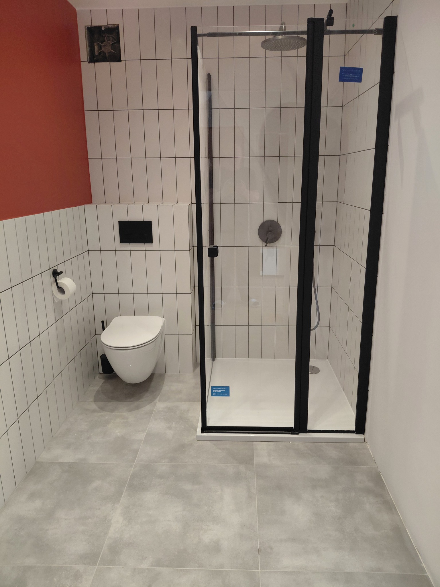 Nowoczesna łazienka z kabiną prysznicową w czarnej ramie, białą wiszącą toaletą i szarą podłogą. Ściany wyłożone białymi, pionowymi płytkami.