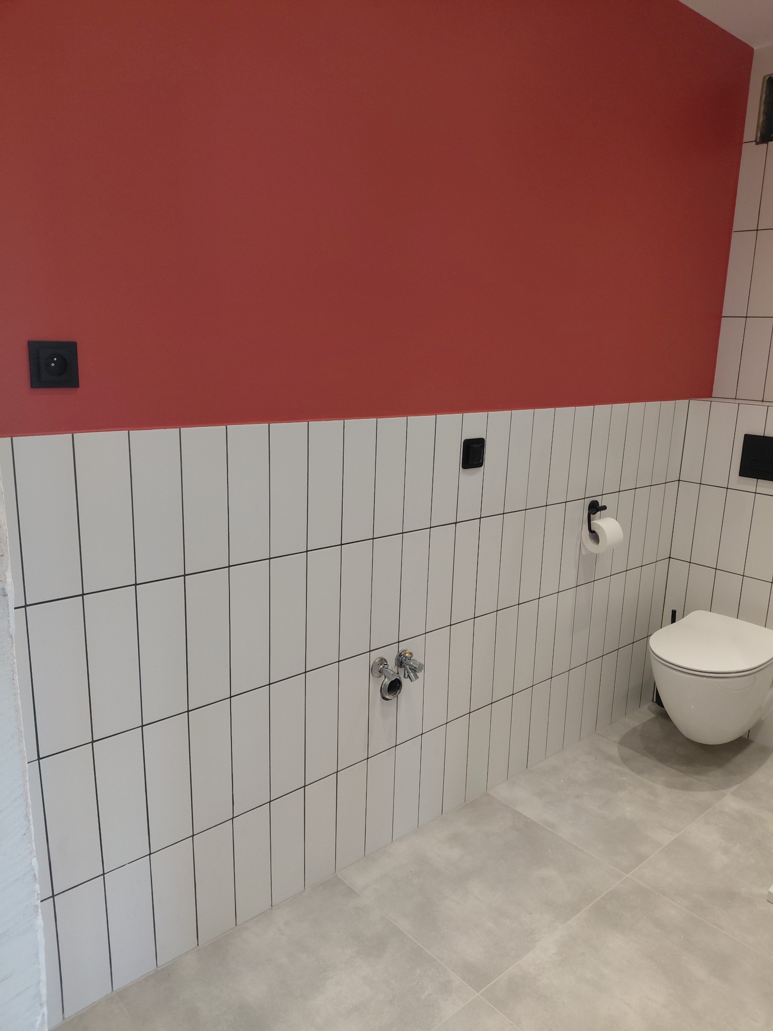 Wykończona łazienka z pionowymi białymi płytkami, czerwoną ścianą, podtynkową toaletą i szarą podłogą. Widoczne czarne gniazdka i uchwyt na papier toaletowy.