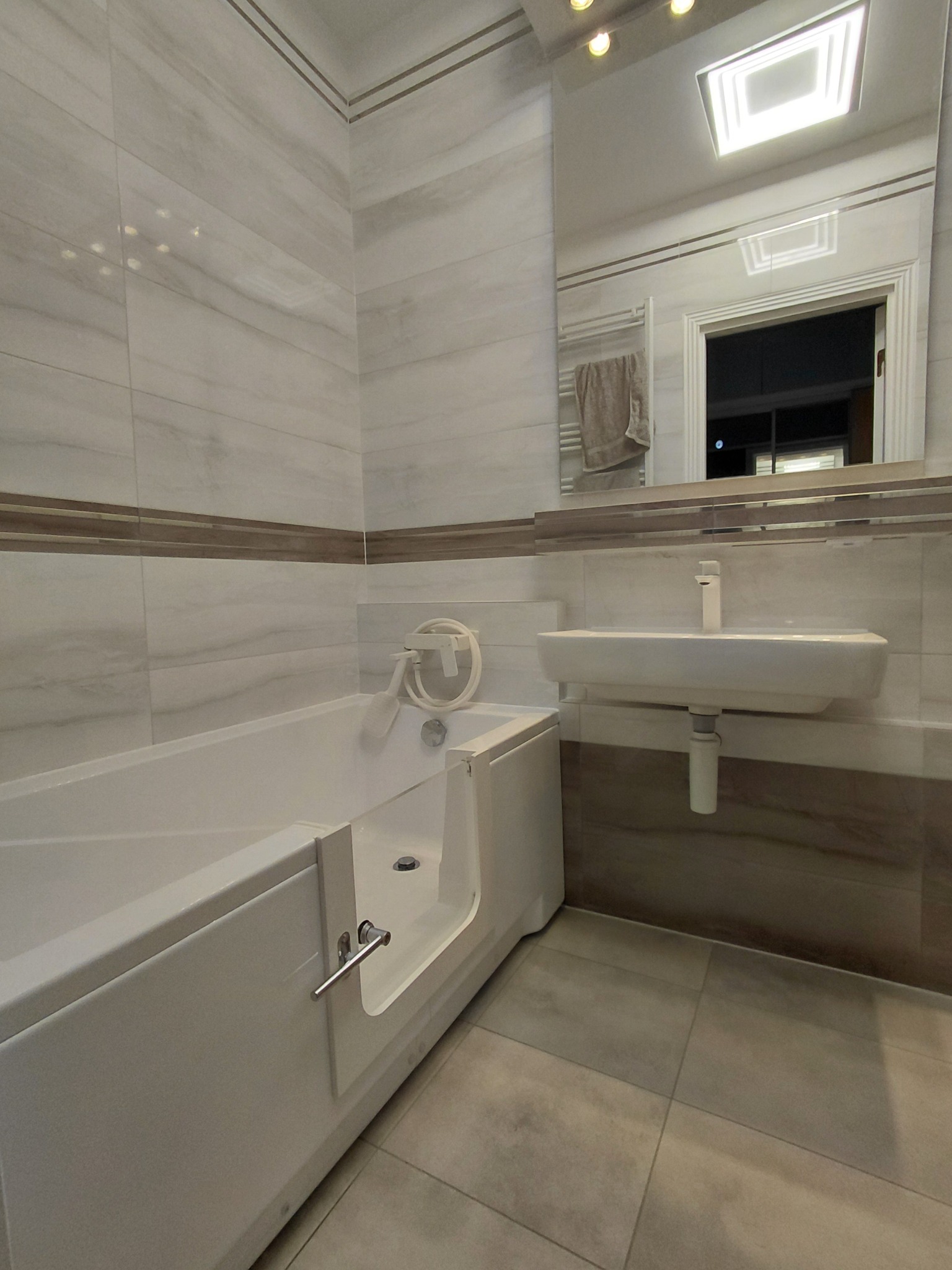 Wykończona łazienka z wanną z drzwiczkami, umywalką i lustrem. Płytki w odcieniach bieli i beżu. Widoczny grzejnik drabinkowy i oświetlenie sufitowe.