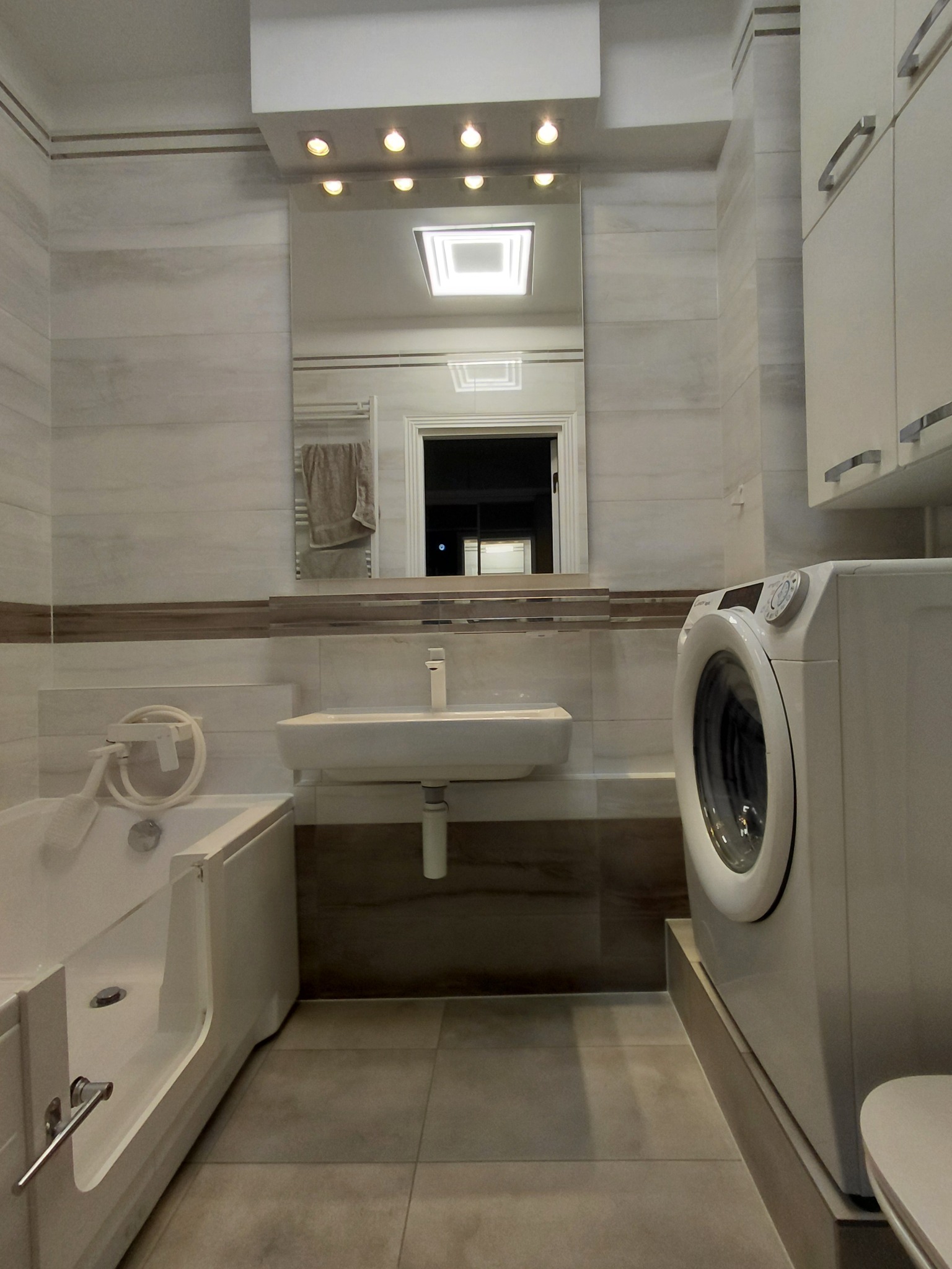 Jasna łazienka z beżowymi płytkami, wanną z otwieranymi drzwiczkami, umywalką i pralką. Lustro z oświetleniem. Minimalistyczny design.