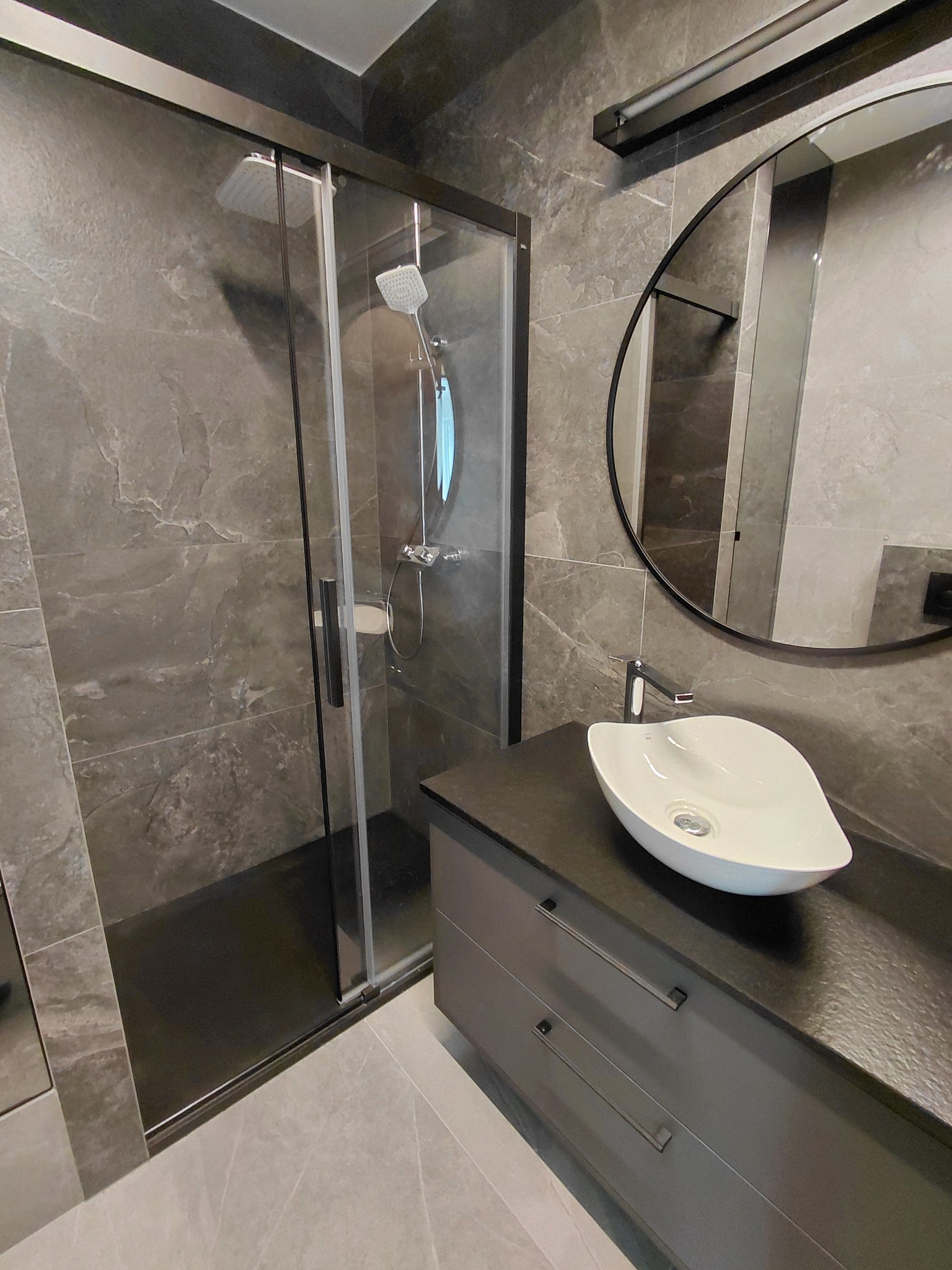 Nowoczesna łazienka w szarościach: kabina prysznicowa z czarnymi ramami, owalna umywalka na szafce z szufladami i okrągłe lustro z czarną ramą.