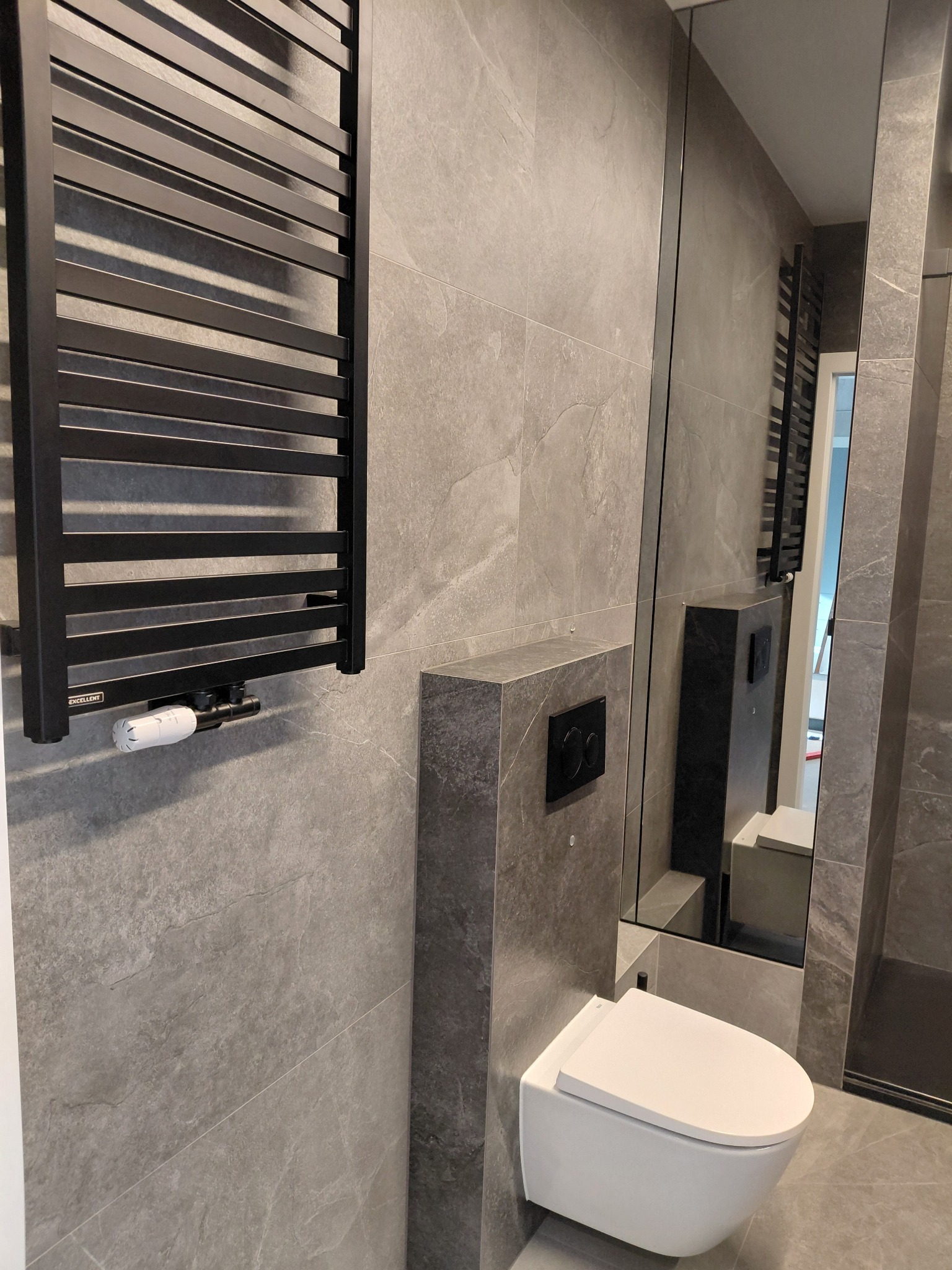 Nowoczesna łazienka w szarościach: wisząca toaleta, grzejnik drabinkowy i duże lustro. Minimalistyczny design z płytkami imitującymi kamień.