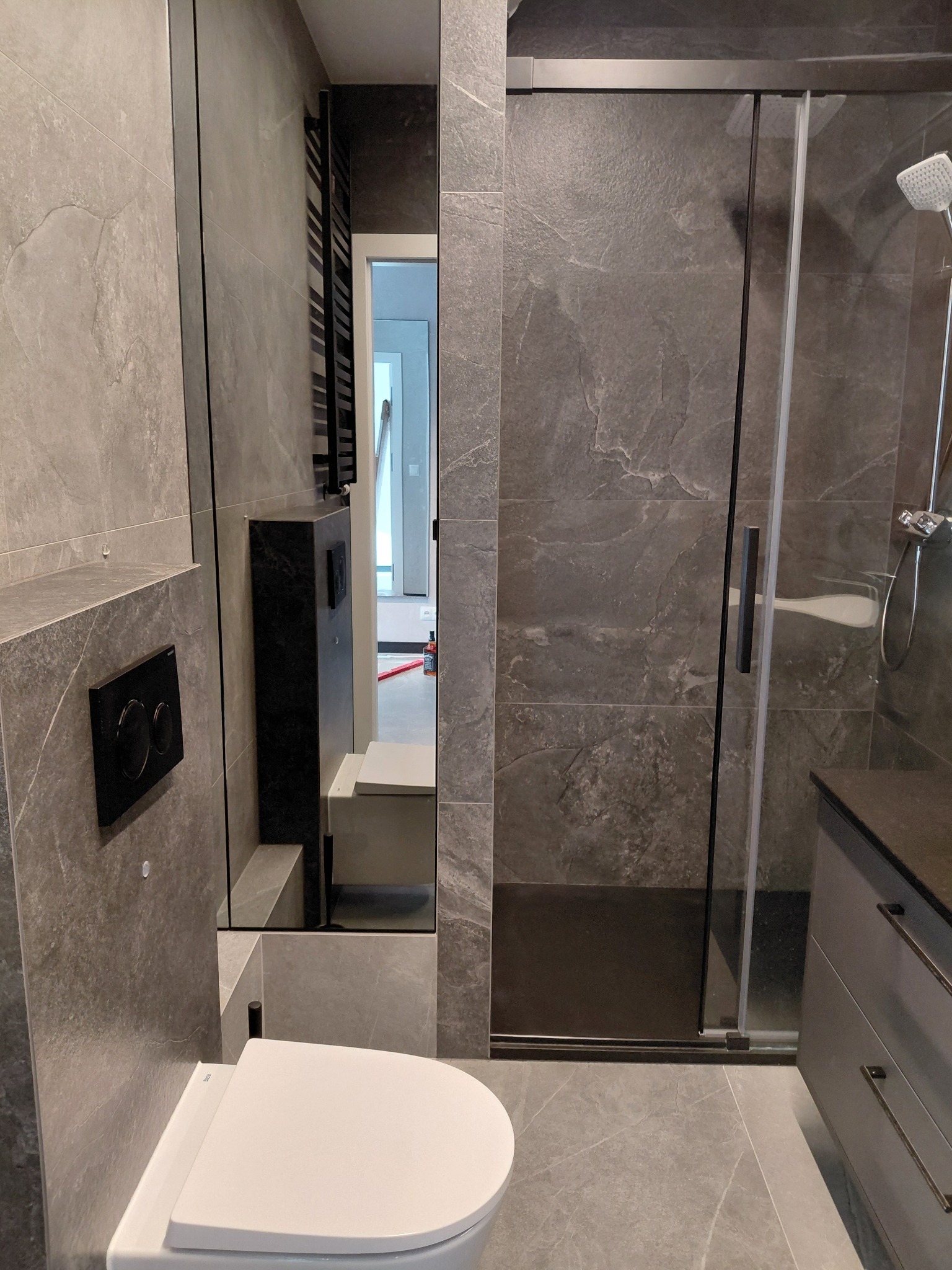 Nowoczesna łazienka w szarościach: kabina prysznicowa ze szklanymi drzwiami, podwieszana toaleta i umywalka z szafką. Minimalistyczny design, płytki imitujące kamień.