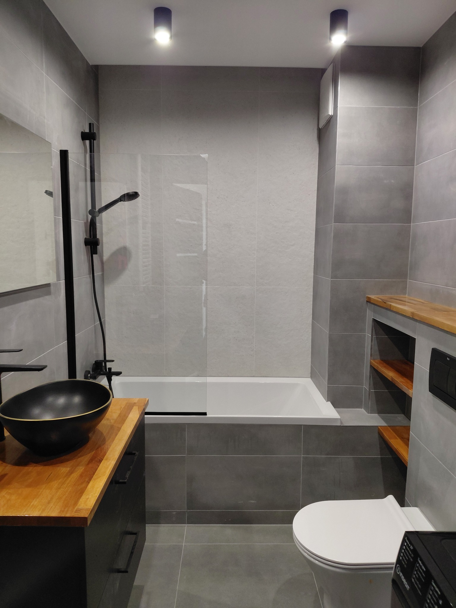 Nowoczesna łazienka w szarościach z drewnianymi akcentami: czarna umywalka nablatowa, bateria i kabina prysznicowa. Minimalistyczny design, płytki imitujące beton.