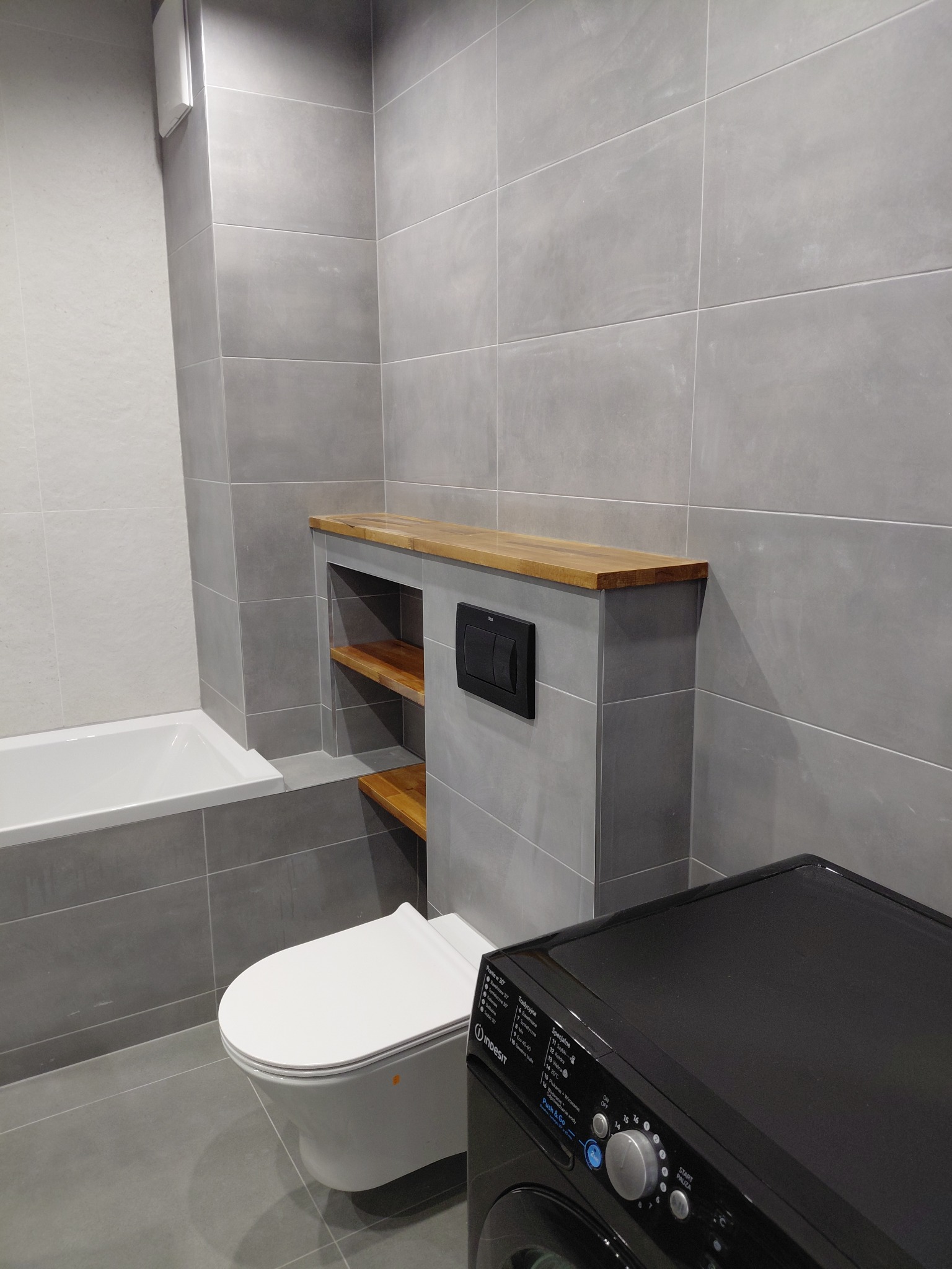 Szara łazienka po remoncie: wisząca toaleta z drewnianą półką, zabudowana spłuczka, pralka Indesit i biała wanna. Minimalistyczny design z płytkami w odcieniach szarości.