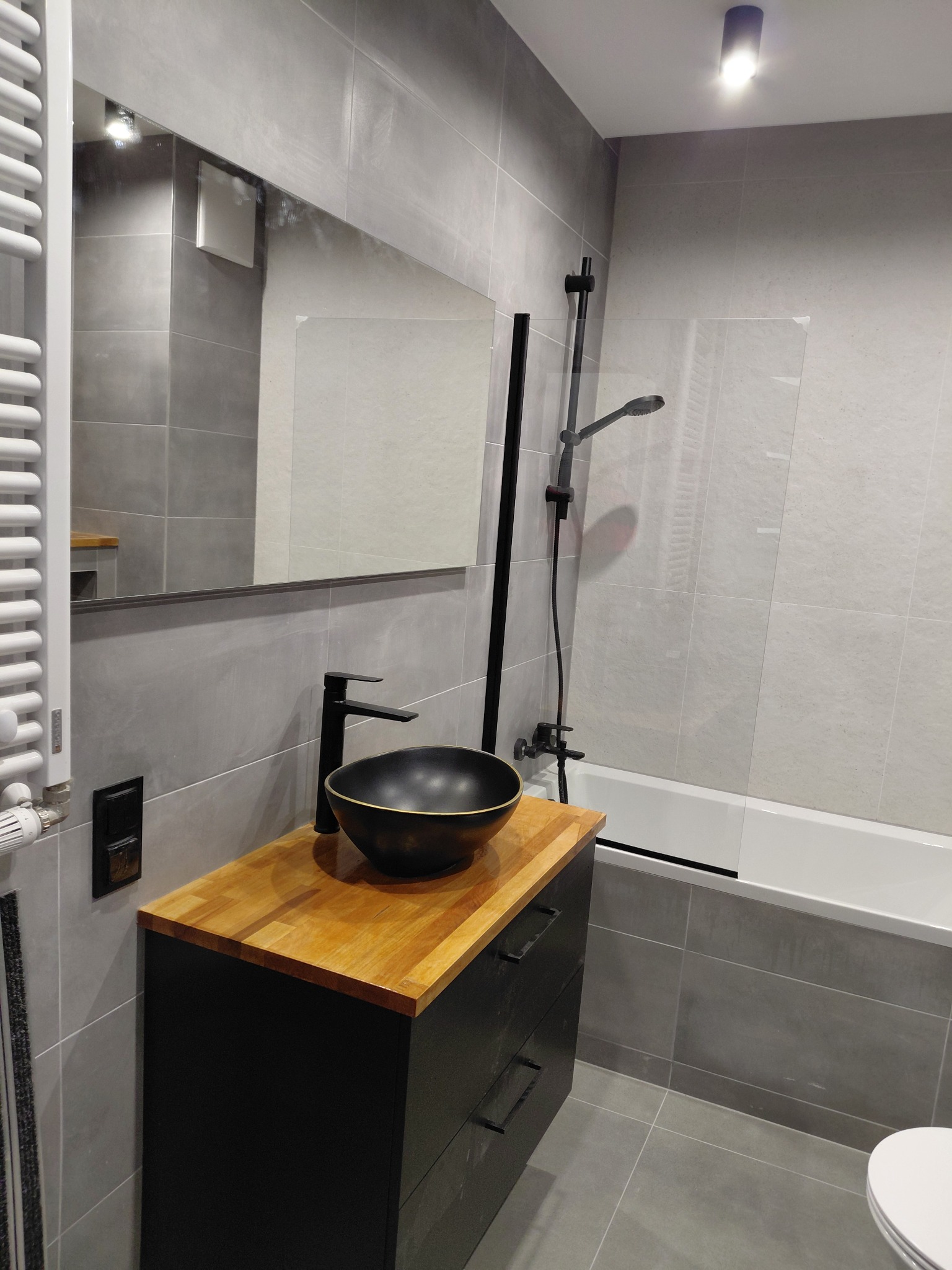 Nowoczesna łazienka w szarościach z drewnianym blatem, czarną umywalką i armaturą. Minimalistyczny design, płytki imitujące beton, grzejnik drabinkowy.