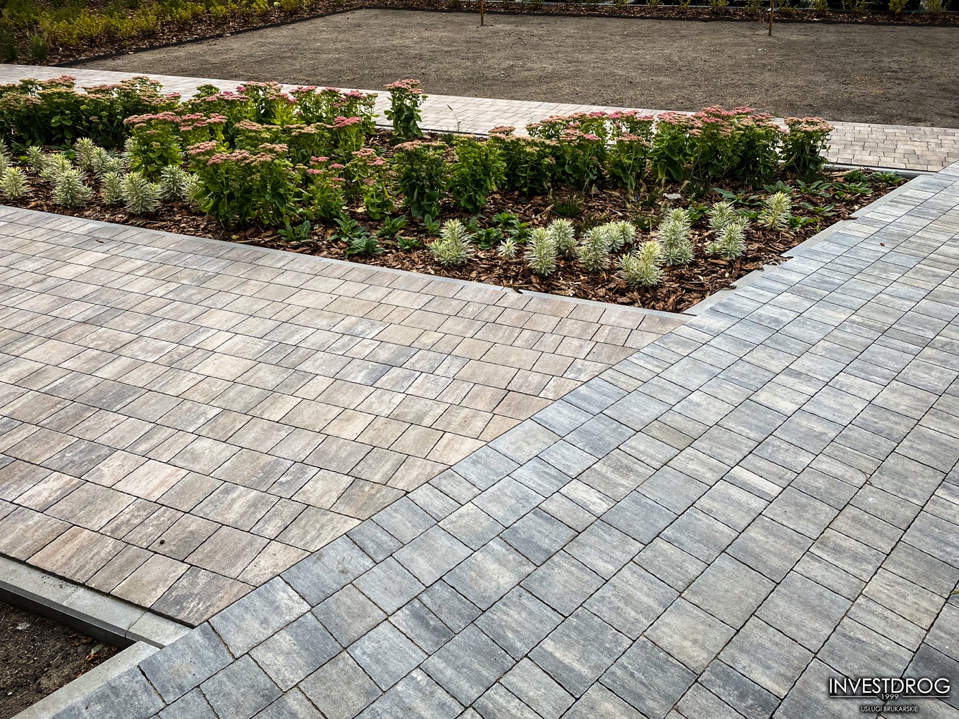 Ułożona kostka brukowa w dwóch odcieniach szarości, tworząca wzór na ścieżce obok rabaty z roślinami kwitnącymi na różowo, widok z góry.