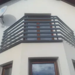 Szara balustrada balkonowa z poziomych profili na rogu budynku, widok z dołu, antena satelitarna w tle.