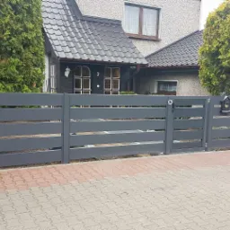 Nowoczesne, poziome ogrodzenie panelowe w kolorze grafitowym z furtką i skrzynką na listy, na tle domu z ciemną dachówką i tujami.