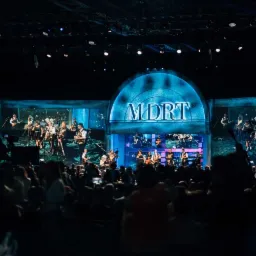 Konferencja MDRT z występem zespołu muzycznego na scenie, widok z perspektywy publiczności, niebieskie oświetlenie sceniczne.