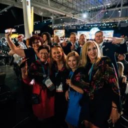 Grupa uśmiechniętych osób pozuje do zdjęcia na tle widowni podczas konferencji, część z nich ma identyfikatory na szyi i tradycyjne chusty w kwiaty.