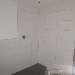 Wnętrze łazienki w trakcie remontu: białe, kwadratowe płytki ścienne z szarą fugą, czarna mozaika podłogowa, drabina malarska i wystający zawór wodny.