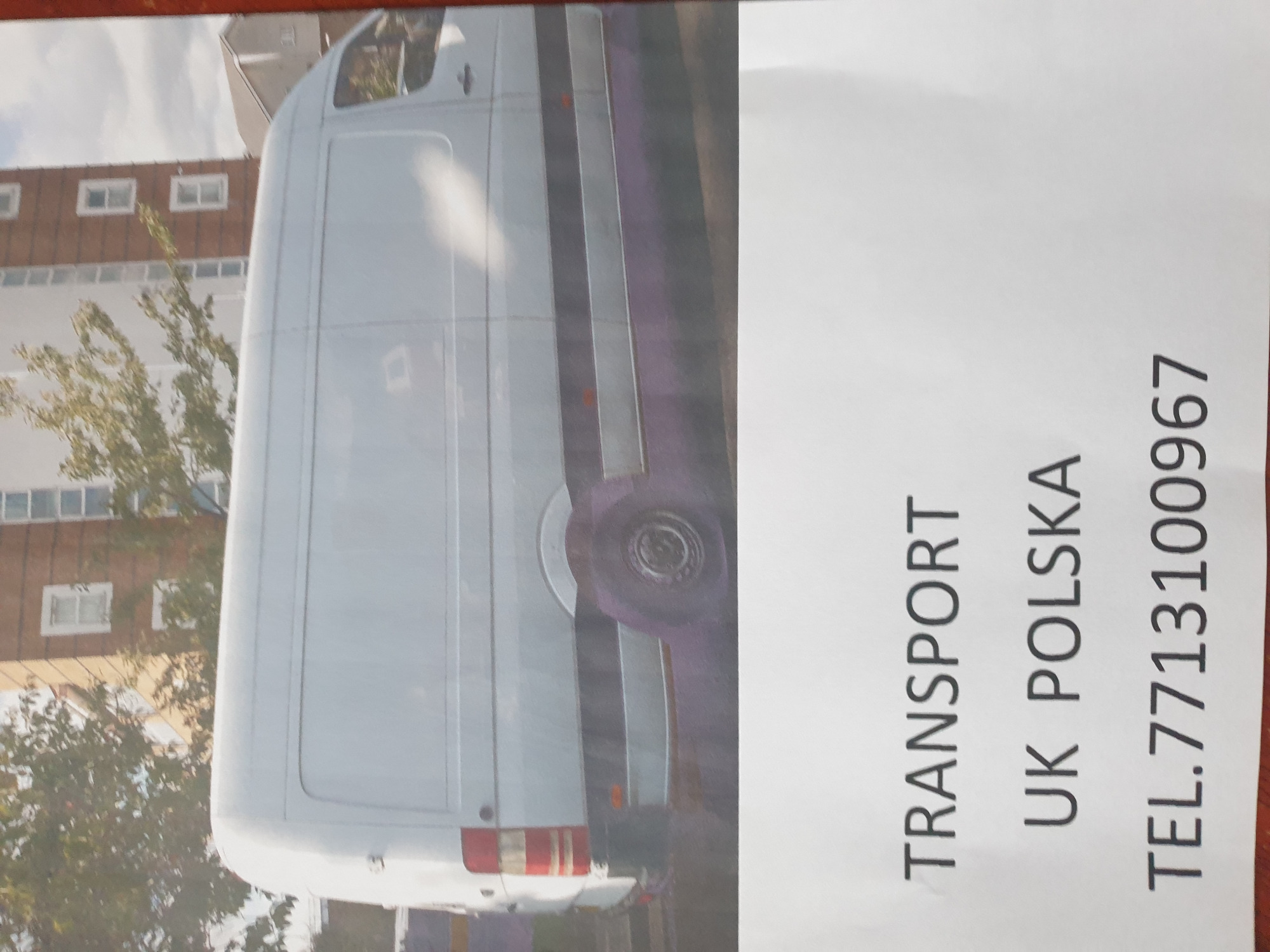 Zdjęcie białego busa na tle budynku mieszkalnego, obok kartka z napisem TRANSPORT UK POLSKA i numerem telefonu